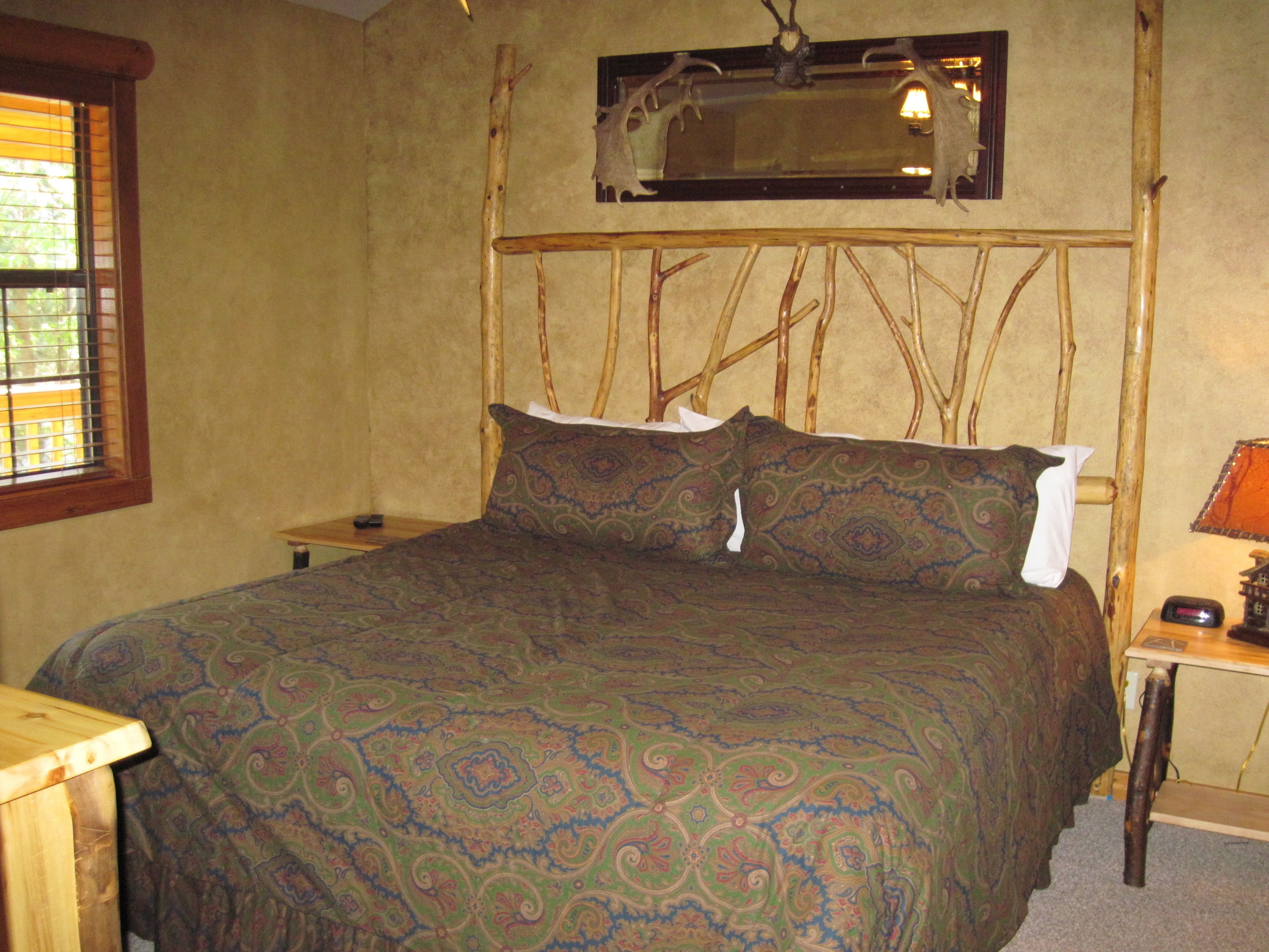A bed with a patterned bedspread, a headboard with branches, a nightstand with a lamp, and a window with blinds.