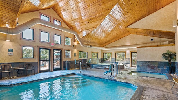 An indoor swimming pool with a wooden ceiling, clear glass doors leading outside, and a seating area with chairs and tables. - The Village At Indian Point Resort (Branson)