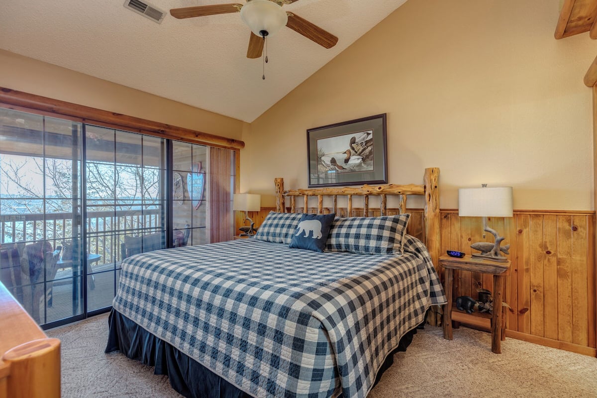 A bedroom with a large bed, a ceiling fan, a wooden headboard, a nightstand, a lamp, and a window with a view of trees.