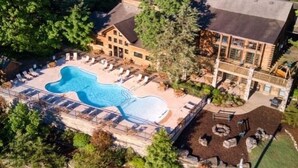 An aerial view of a resort with a large swimming pool surrounded by lounge chairs, a building with multiple windows, and a forested area. - The Village At Indian Point Resort (Branson)