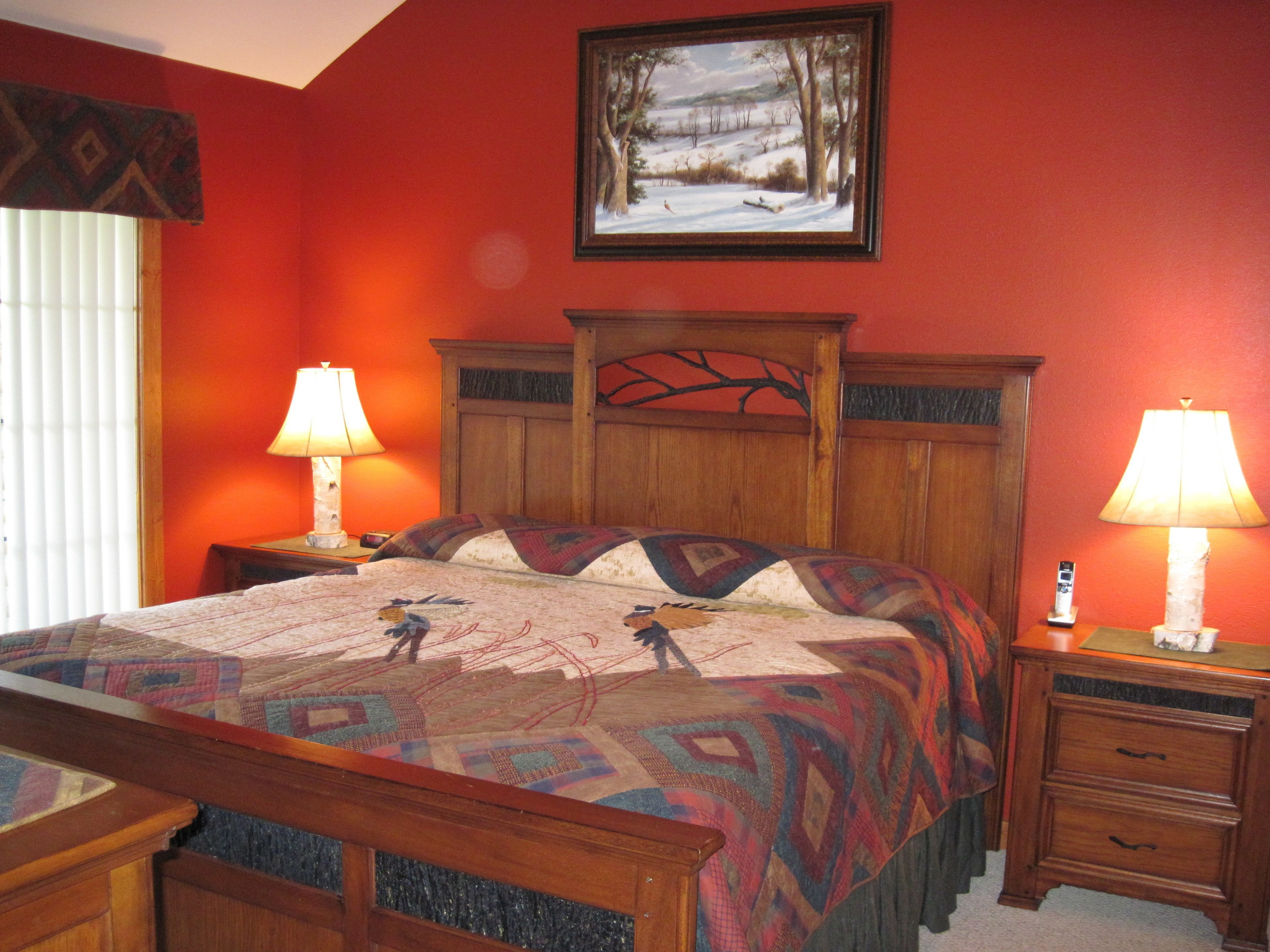 A bedroom with a wooden headboard, a quilt with a bird design, two bedside tables with lamps, and a painting of a snowy landscape on the wall.