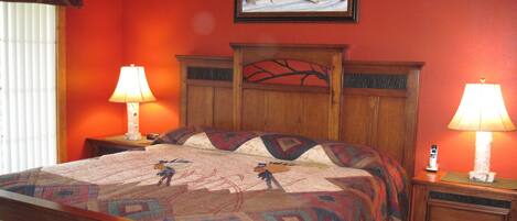 A bedroom with a wooden headboard, a quilt with a bird design, two bedside tables with lamps, and a painting of a snowy landscape on the wall.