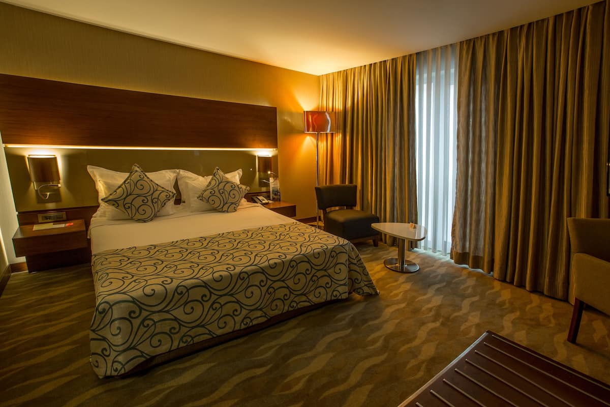 A hotel room with a large bed, a bedside table with a lamp, a chair, a small table, and a window with curtains.