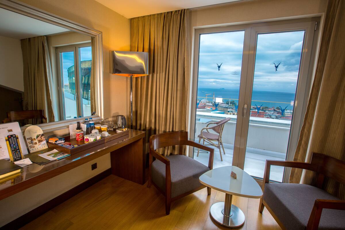 A hotel room with a desk, two chairs, a small table, a TV, and a large window with a view of the sea and buildings.