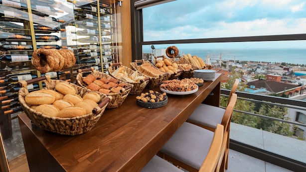 Une table de buffet garnie de différents pains et pâtisseries, un présentoir à vin en arrière-plan et une vue sur la mer par une grande fenêtre.