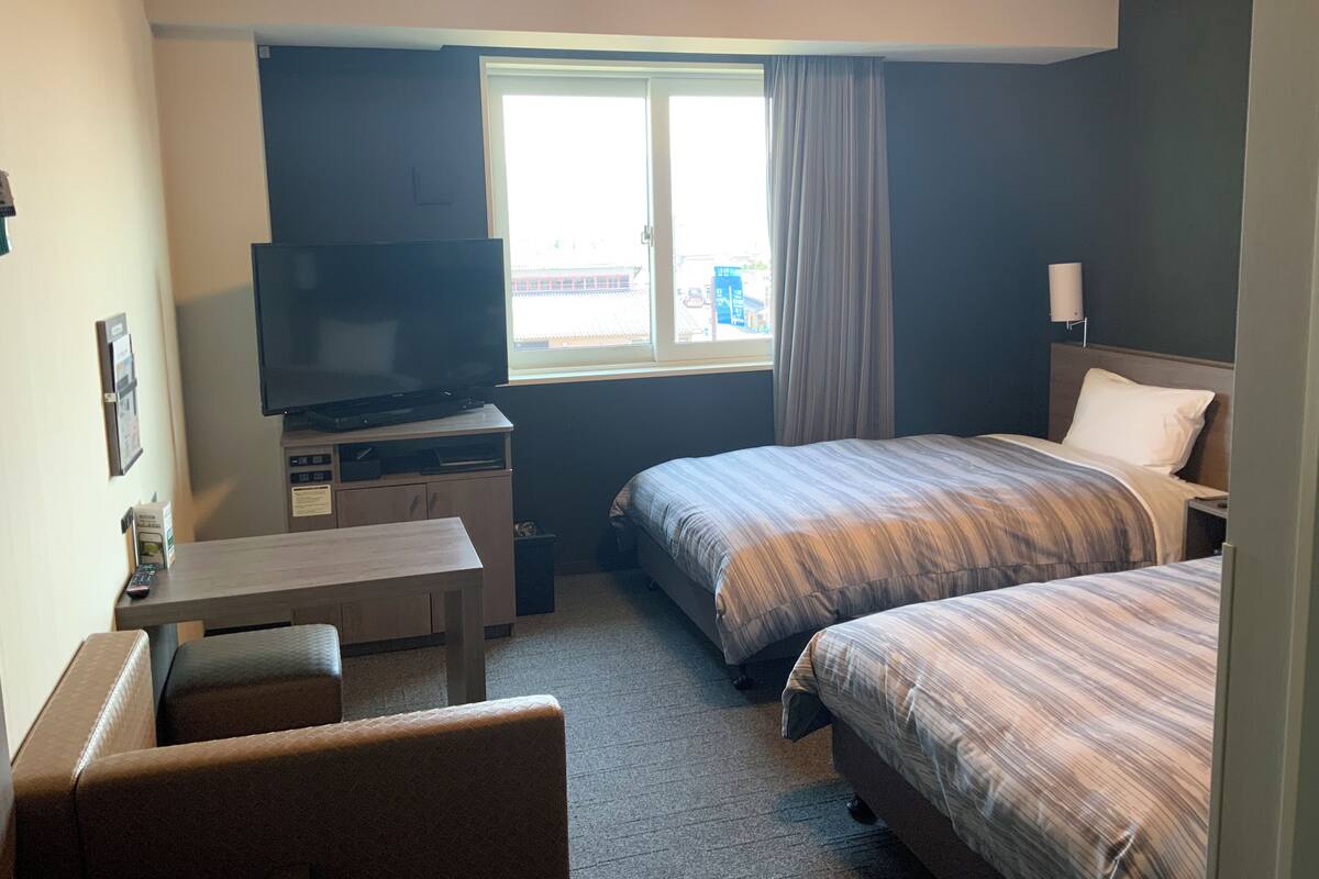 A hotel room with a TV, two beds, a desk, and a window with curtains.