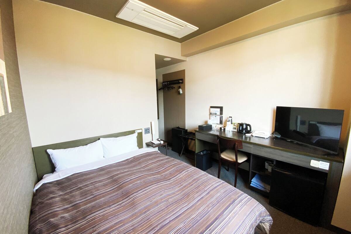 A hotel room with a bed, desk, chair, TV, and a trash bin.