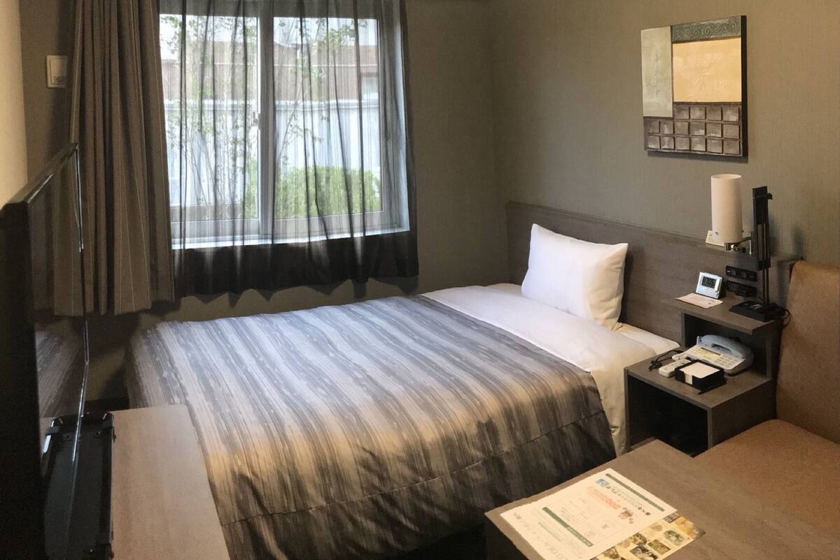 A hotel room with a bed, a desk, a chair, a nightstand, a lamp, a clock, and a window with curtains.