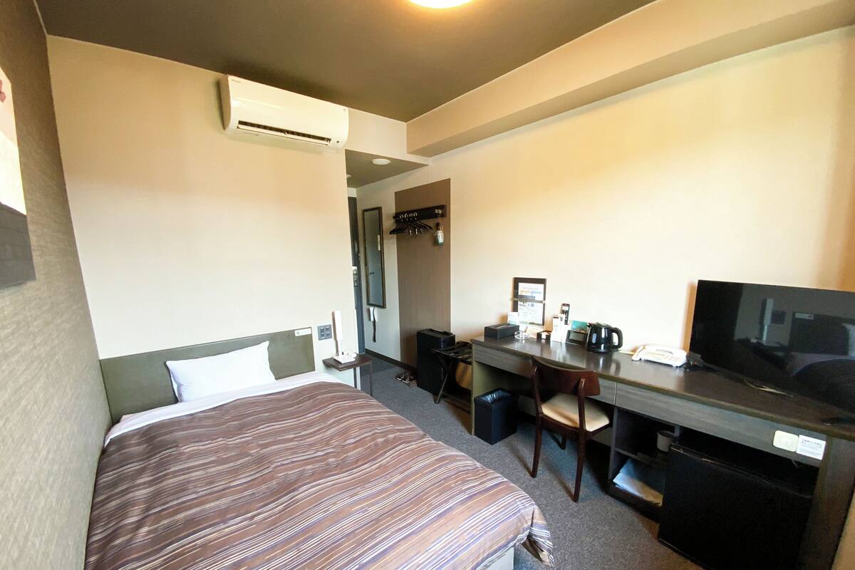 A hotel room with a bed, desk, chair, TV, and a small trash bin.