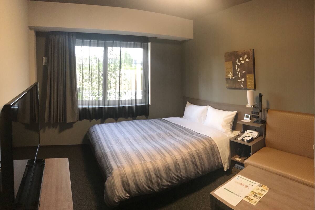 A hotel room with a bed, a desk, a chair, a window with curtains, and a wall-mounted lamp.
