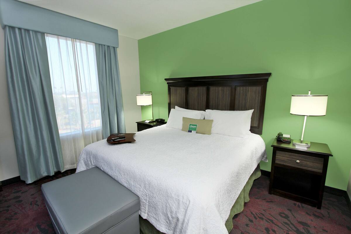 A hotel room with a large bed, two bedside tables with lamps, a window with curtains, and a small bench.