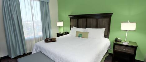 A hotel room with a large bed, two bedside tables with lamps, a window with curtains, and a small bench.