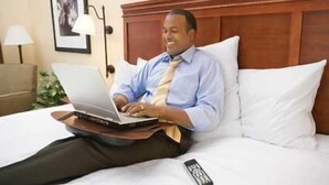 A man sitting on a bed with a laptop, a remote control, and a framed picture on the wall.