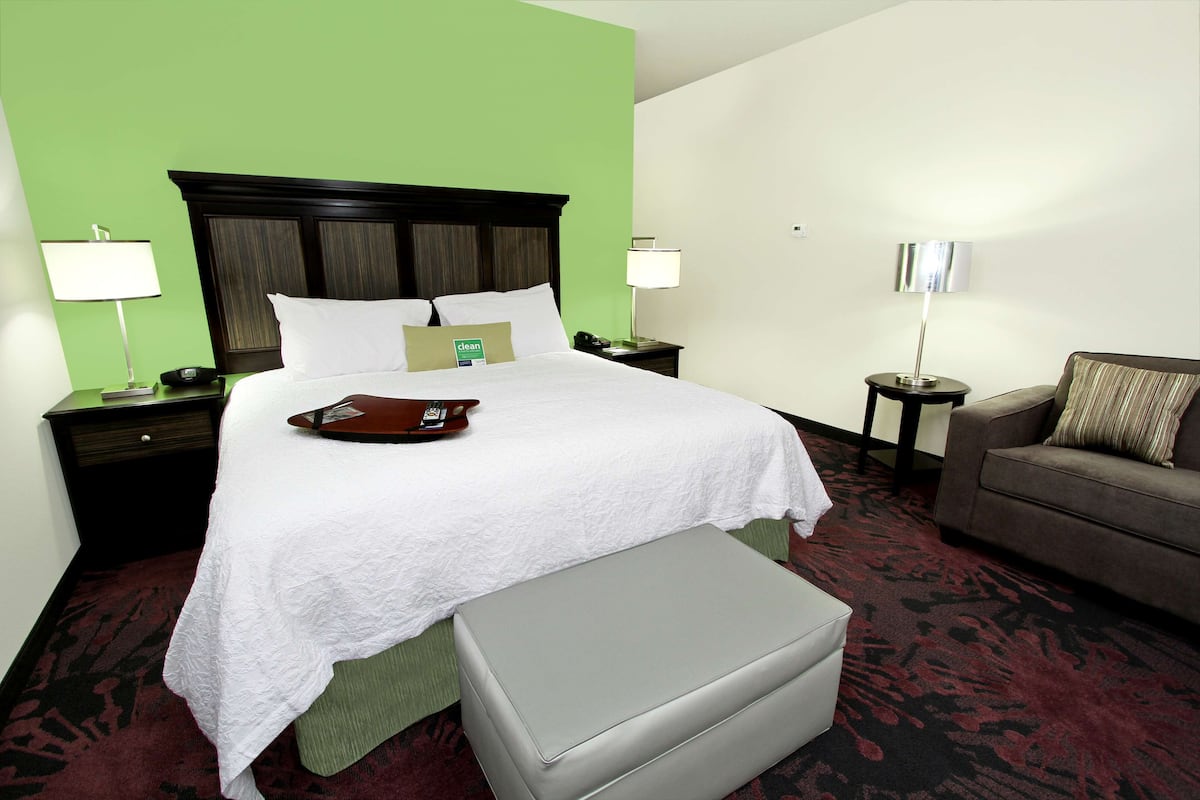 A hotel room with a large bed, a grey ottoman, a brown armchair, and two bedside tables with lamps.