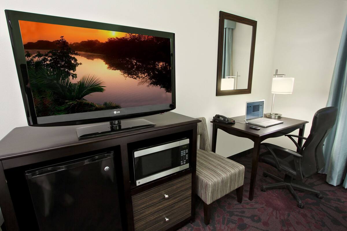A hotel room with a flat-screen TV, a desk with a laptop, a chair, a microwave, and a small refrigerator.