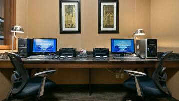 A hotel workspace with two desks, each with a desktop computer, keyboard, mouse, and monitor. There are two framed pictures on the wall above the desks.