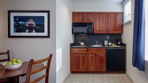 A small kitchen with wooden cabinets, a black microwave, and a sink. There is a bowl of fruit on the table and a chair with a red cushion. - Federal City Inn & Suites (New Orleans)
