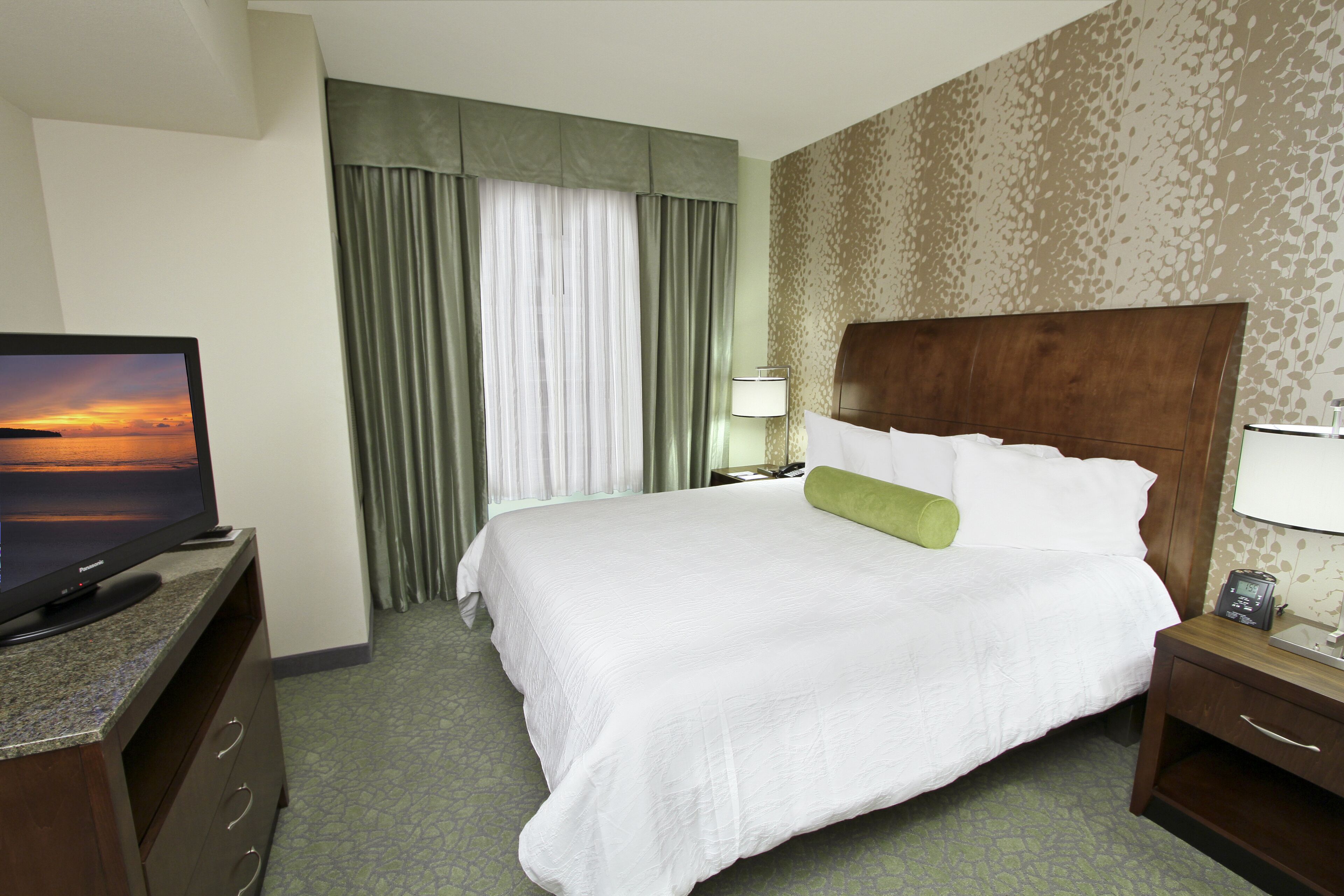 A hotel room with a large bed, a television on a dresser, a nightstand with a lamp, and a window with curtains.