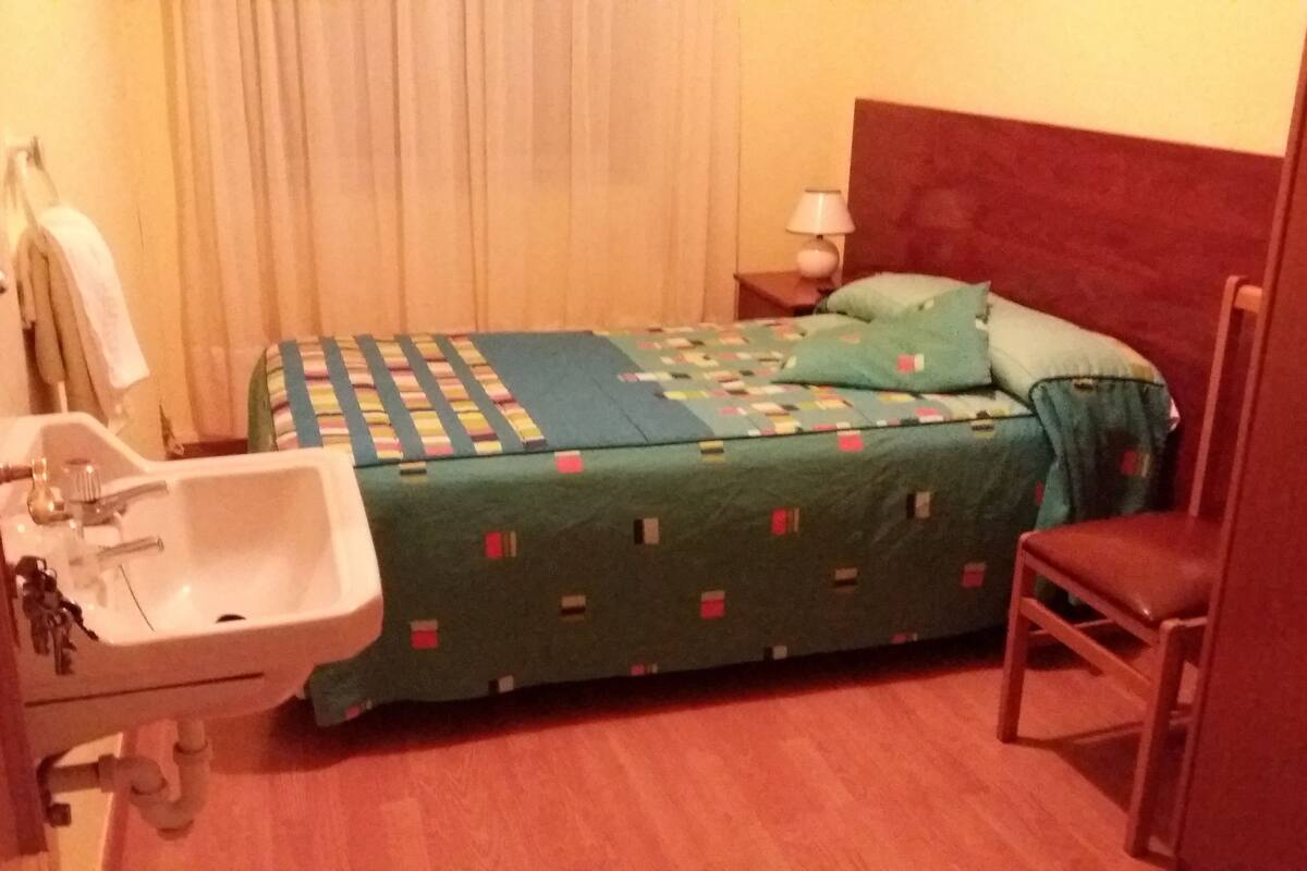 A small hotel room with a single bed, a bedside table with a lamp, a chair, a sink, and a mirror.