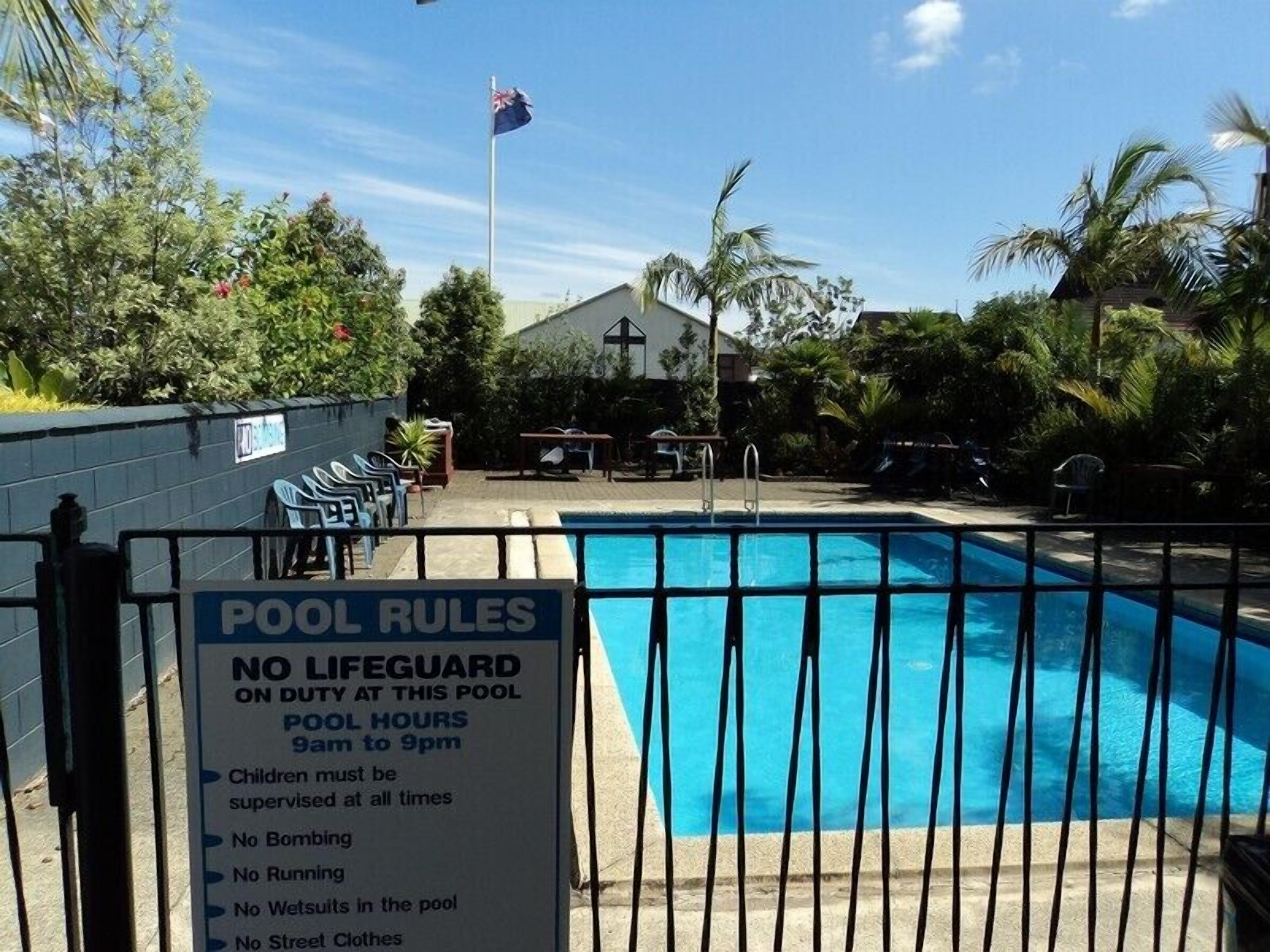 A pool with a sign listing rules such as no lifeguard on duty, pool hours from 9am to 9pm, and restrictions like no running, no wet suits, and no street clothes.
