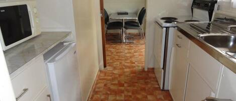 Cocina compacta con microondas, estufa y lavaplatos. Se ve un área de comedor con una mesa y sillas al fondo.