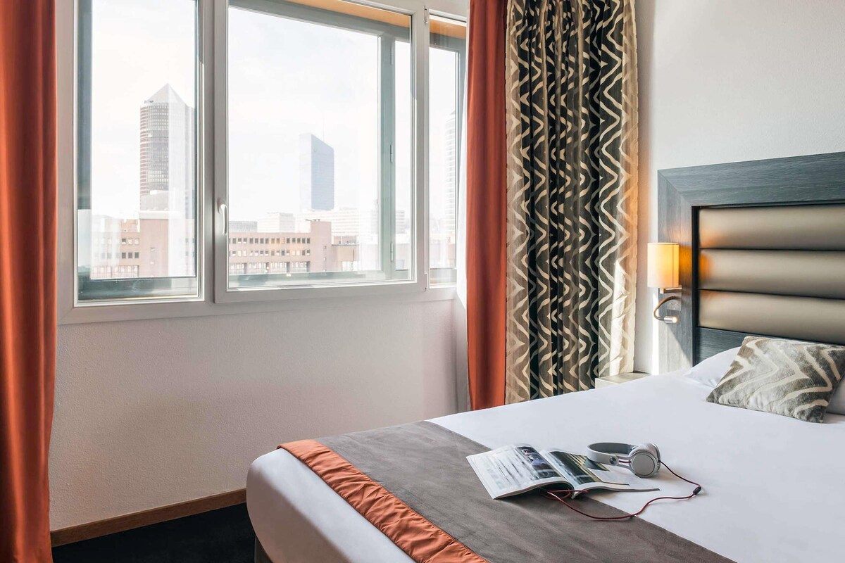 Une chambre d’hôtel avec un lit, une fenêtre avec des rideaux, une vue sur la ville et une tête de lit dotée d’une lampe.