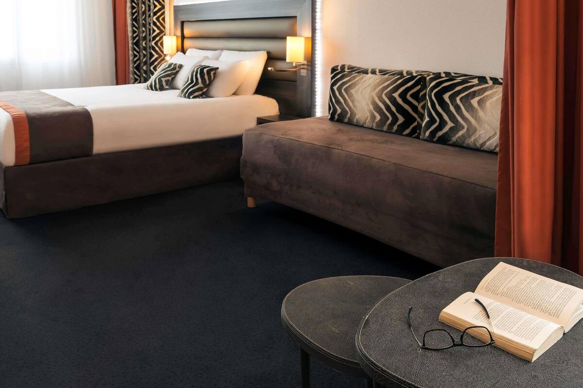 A hotel room with a bed, a sofa, and a small table with a book and glasses.