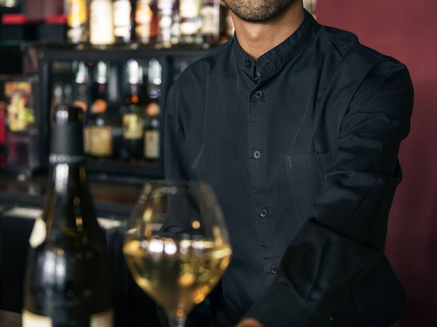 A person in a black shirt standing behind a bar with a bottle of wine and a glass.