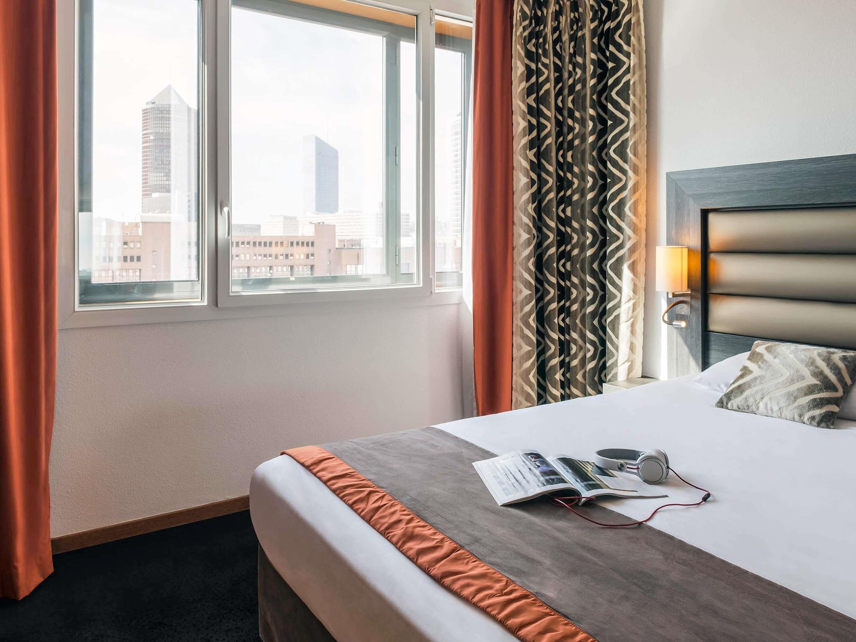 Ein Hotelzimmer mit Bett, Vorhängen am Fenster, Blick auf die Stadt und einer Kopfteil mit Lampe.