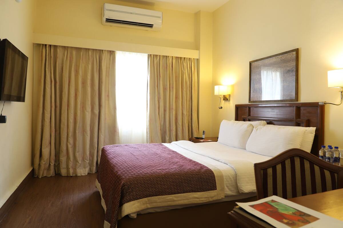 A hotel room with a bed, a desk, a chair, a television, a painting, and a window with curtains.