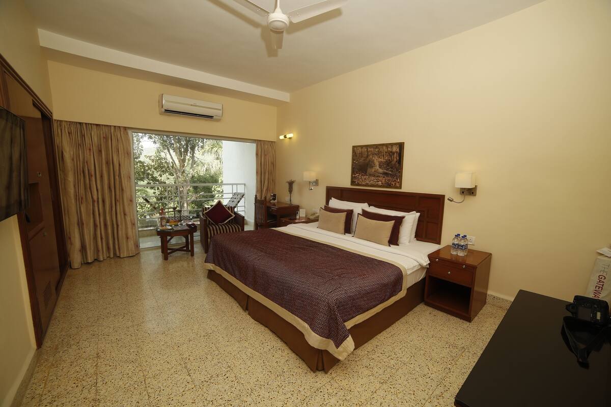 A hotel room with a large bed, a nightstand, a desk, and a chair. There is a window with curtains, a ceiling fan, and an air conditioning unit.