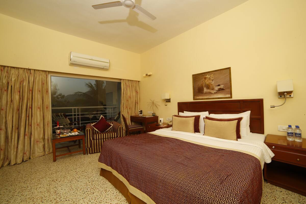 A hotel room with a large bed, a desk, a chair, a window with curtains, a ceiling fan, an air conditioning unit, a painting, and a nightstand with water bottles.