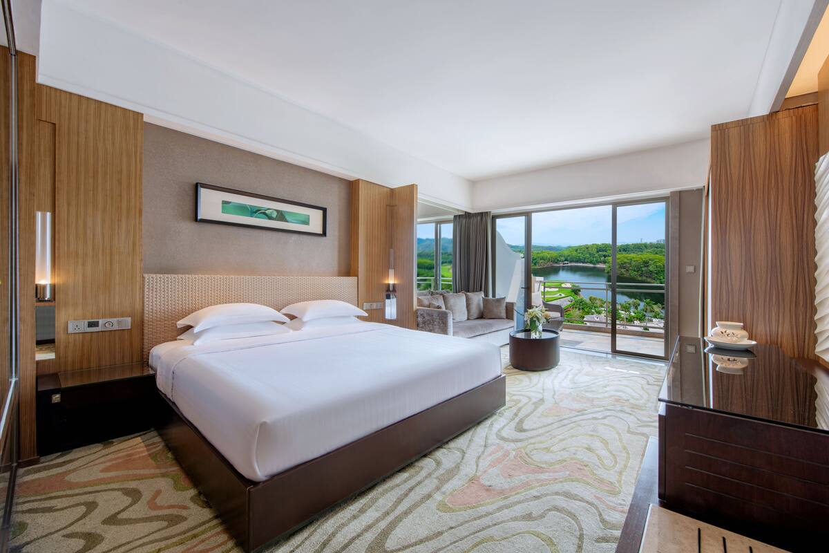 Una habitación de hotel moderna con una cama grande, un sofá, una mesa de comedor y vistas a un paisaje verde a través de la puerta de cristal.