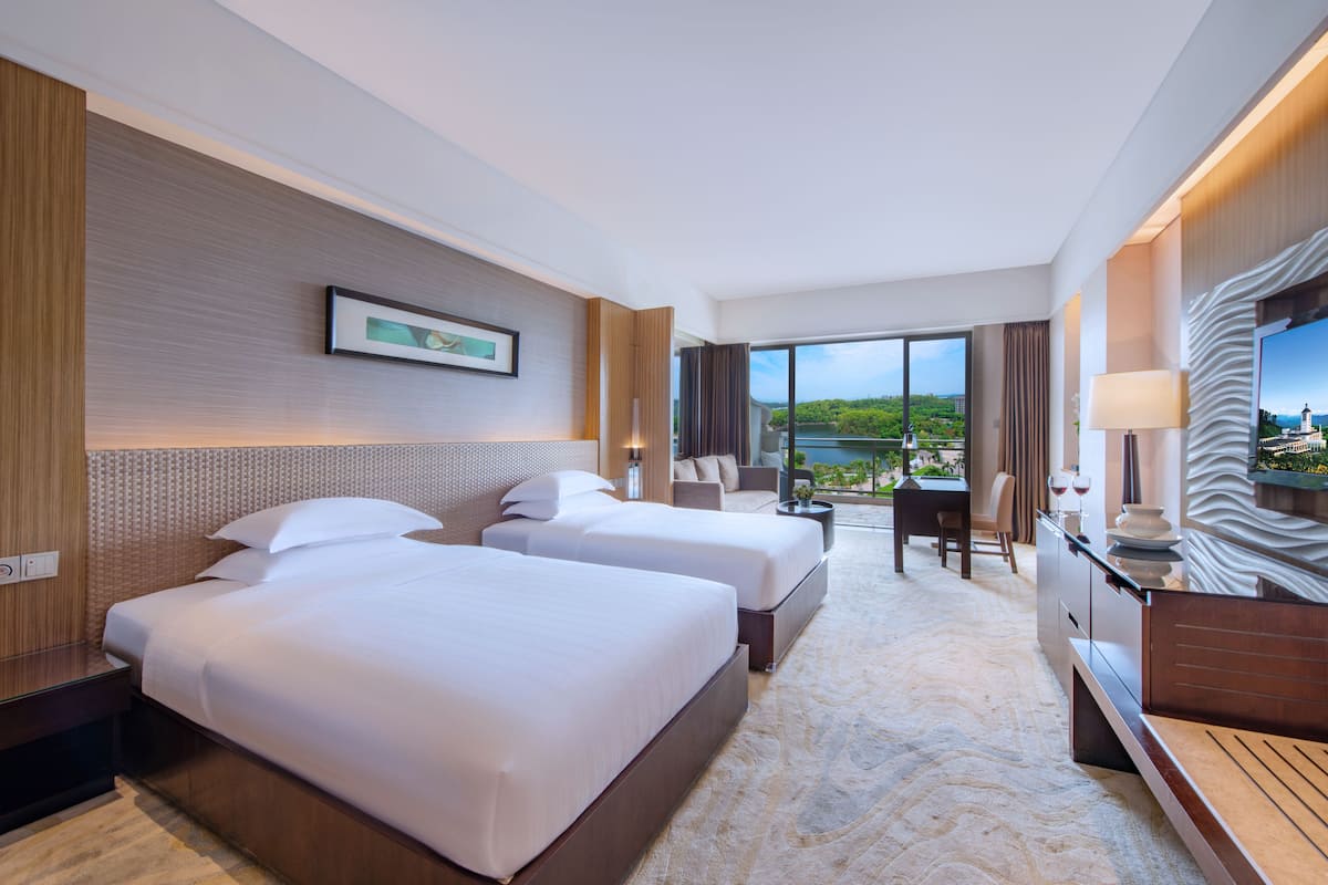 Habitación de hotel con dos camas, un escritorio, una silla, un televisor y un amplio ventanal con vistas a zonas verdes.