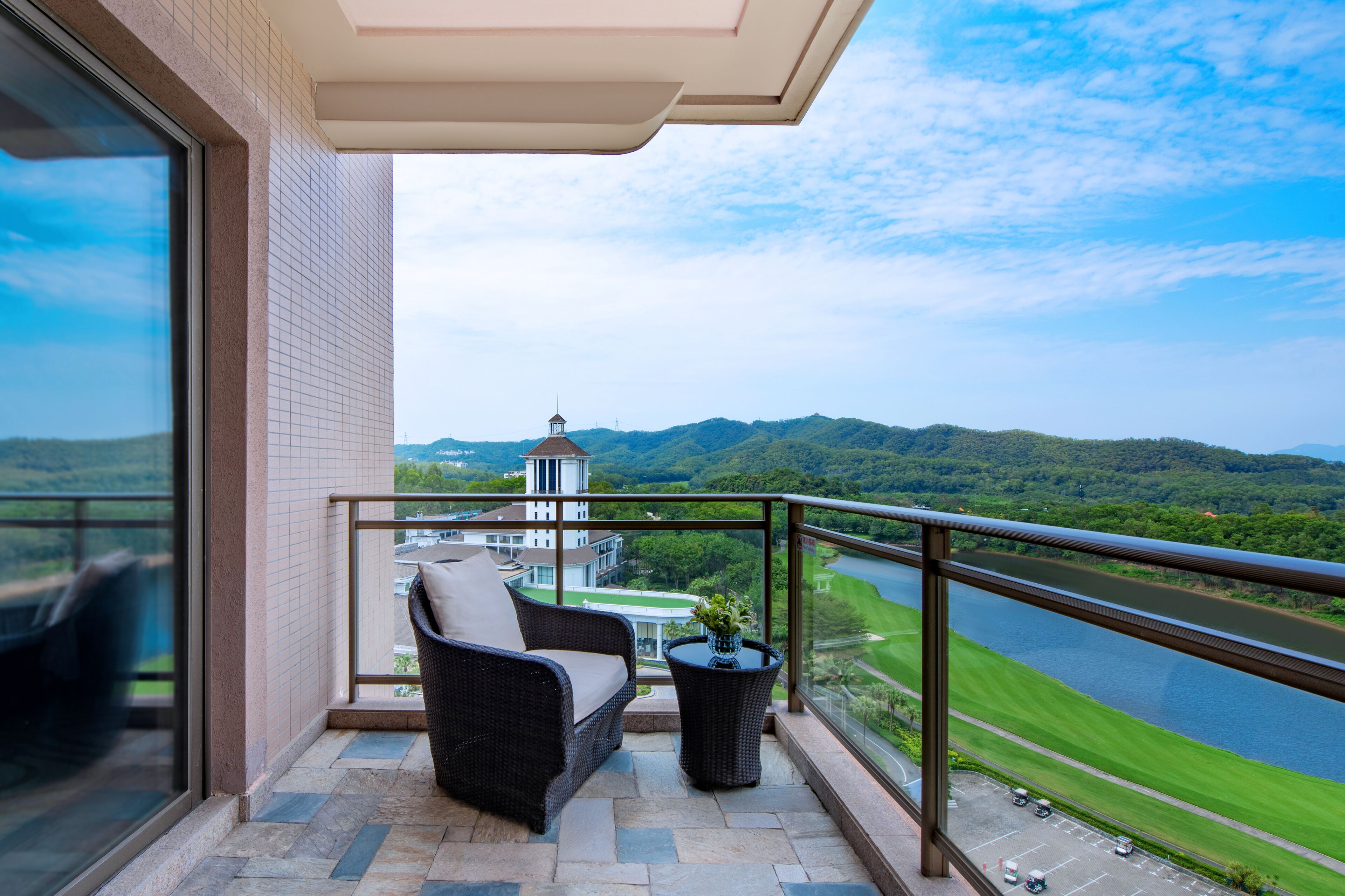 A balcony with a wicker sofa and a small table, overlooking a river and green hills.