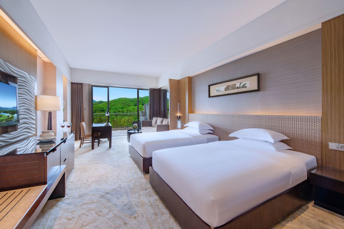 Habitación de hotel con dos camas, un escritorio, una silla y un amplio ventanal con vistas a zonas verdes.
