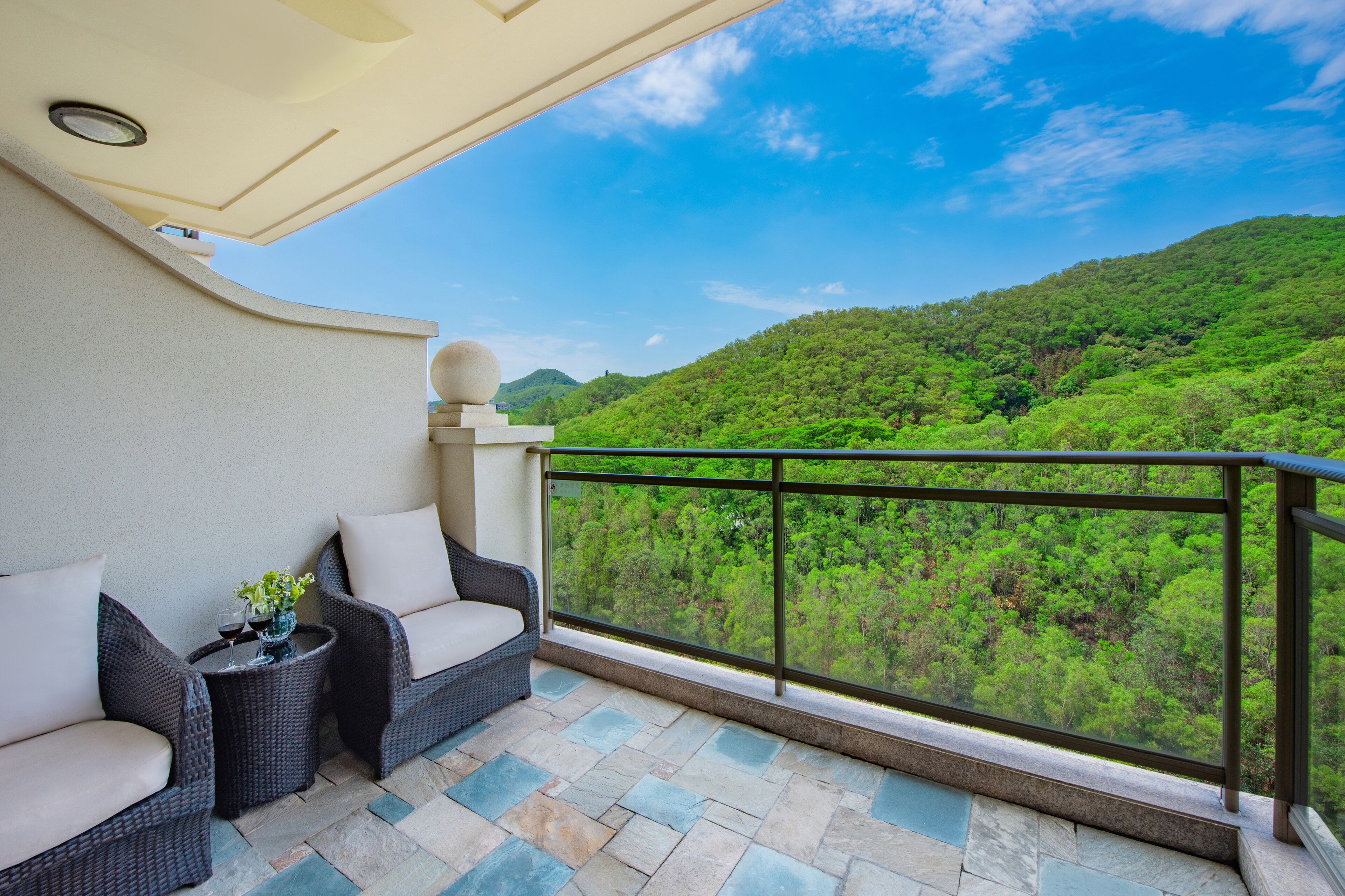 A balcony with wicker furniture, a glass railing, and a view of a lush green hillside.