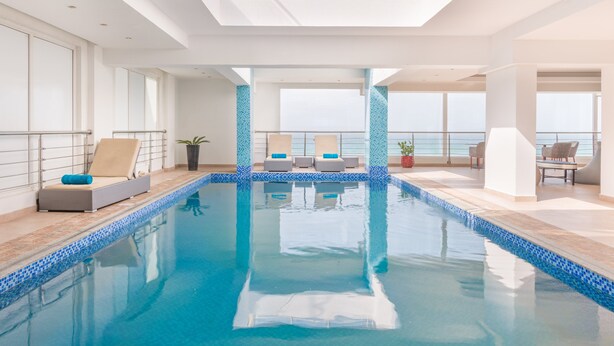 A modern indoor swimming pool with clear blue water, sun loungers, and a view of the sea. - Al Hail Waves Hotel Managed By Centara (Seeb)