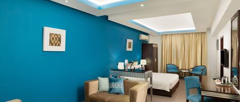 A hotel room with a blue accent wall, a sofa, a dining table, a bed, and a TV.