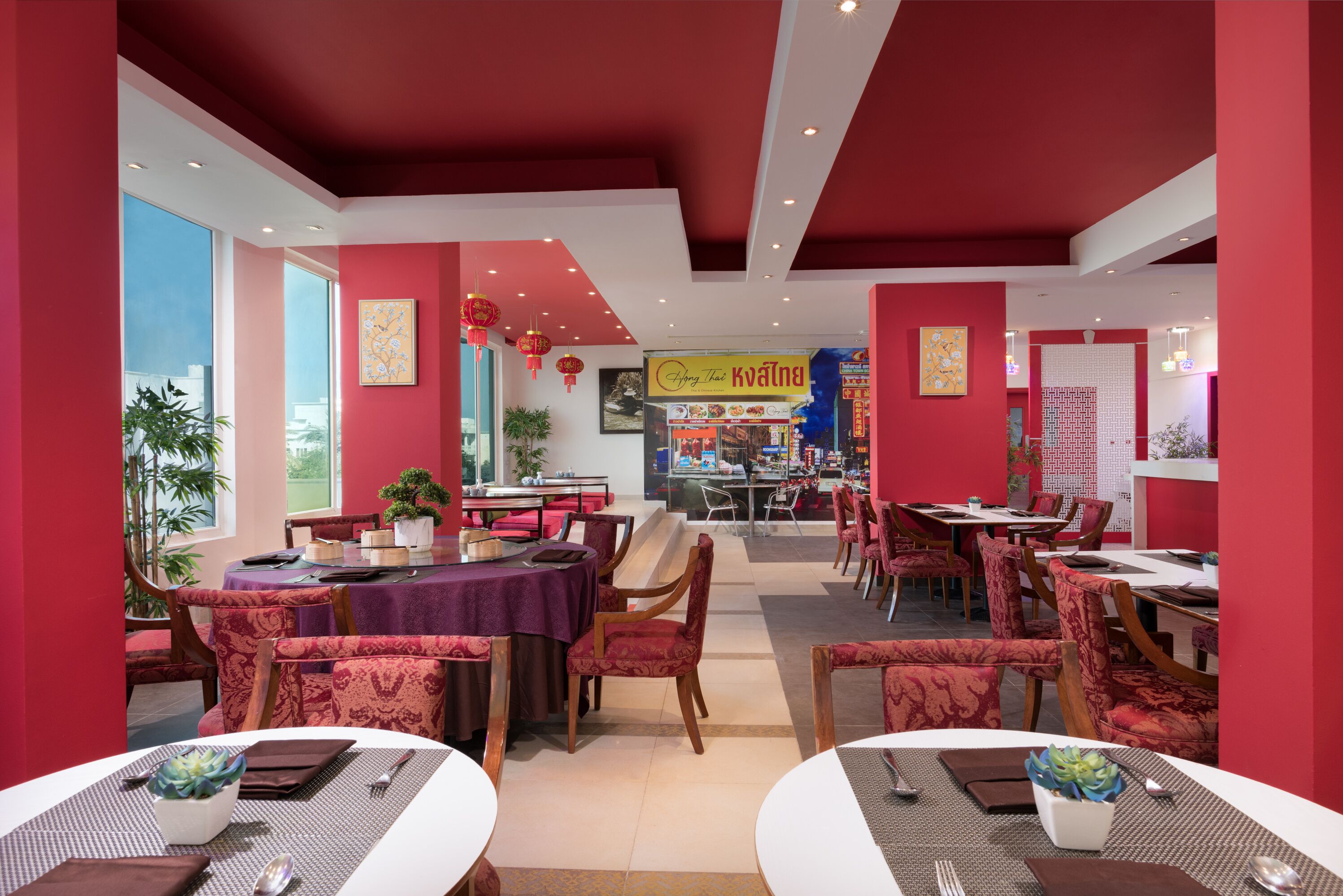 A restaurant with red walls, multiple dining tables, and chairs. There are potted plants and a sign with Thai food.