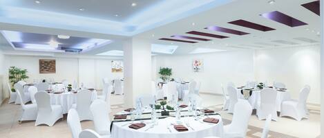A spacious banquet hall with round tables set for dining, white chairs, and ceiling lights.