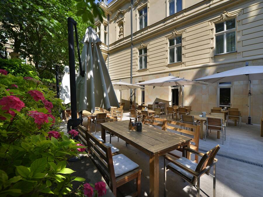 An outdoor cafe area with wooden tables and chairs, surrounded by lush greenery and pink flowers.