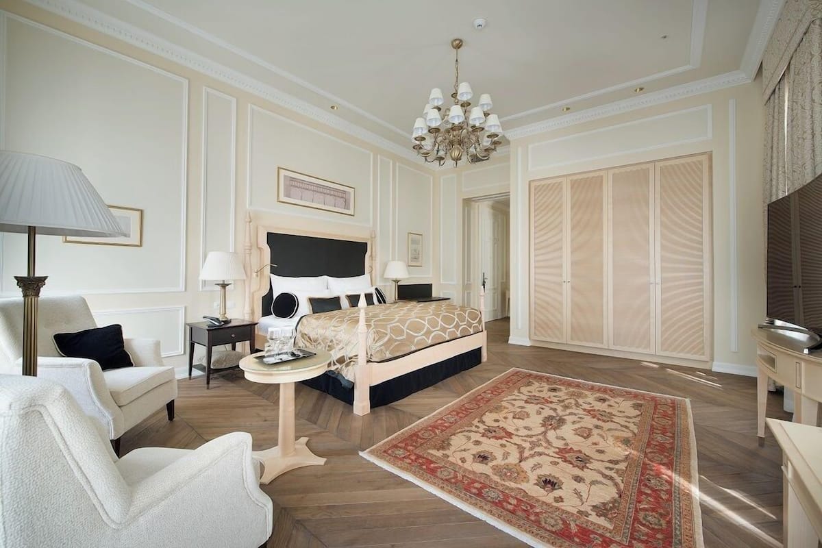 A spacious bedroom with a large bed, a chandelier, a bedside table, a nightstand, a chair, a sofa, and a wardrobe.