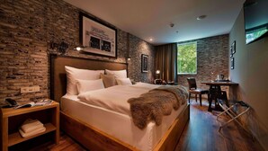 Una habitación de hotel moderna con pared de ladrillo, una cama grande con ropa blanca, un escritorio de madera y vistas a zonas verdes.
