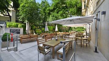 An outdoor cafe with wooden tables and benches, umbrellas, and a stone wall.