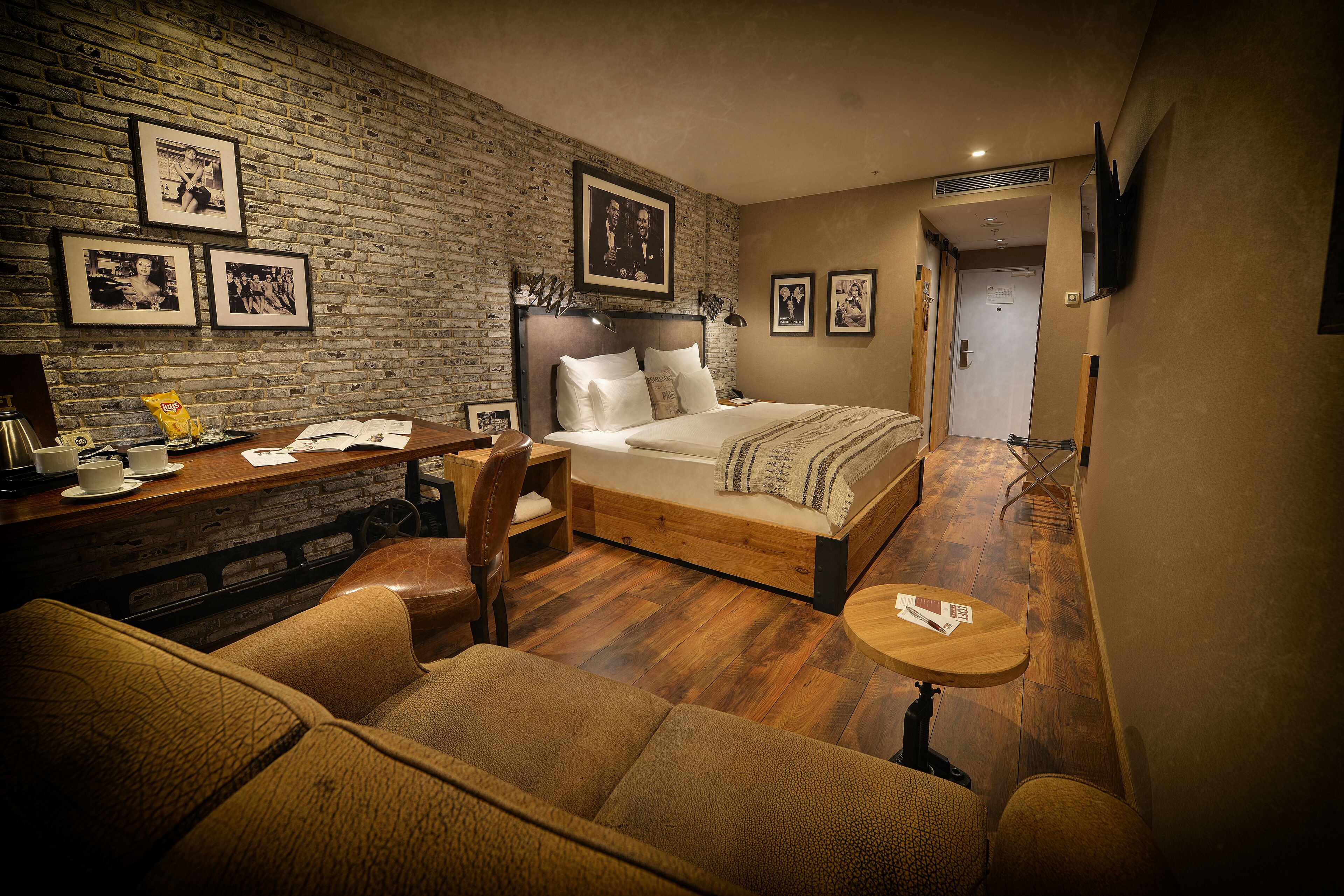 A cozy hotel room with a brick wall, a bed with a wooden headboard, a small desk, a sofa, and a TV mounted on the wall.