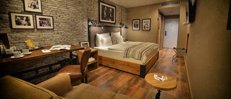A cozy hotel room with a brick wall, a bed with a wooden headboard, a small desk, a sofa, and a TV mounted on the wall.