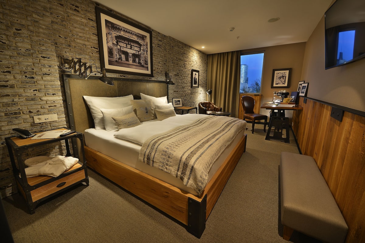 A hotel room with a brick wall, a wooden bed, a desk, and a view of a cityscape.