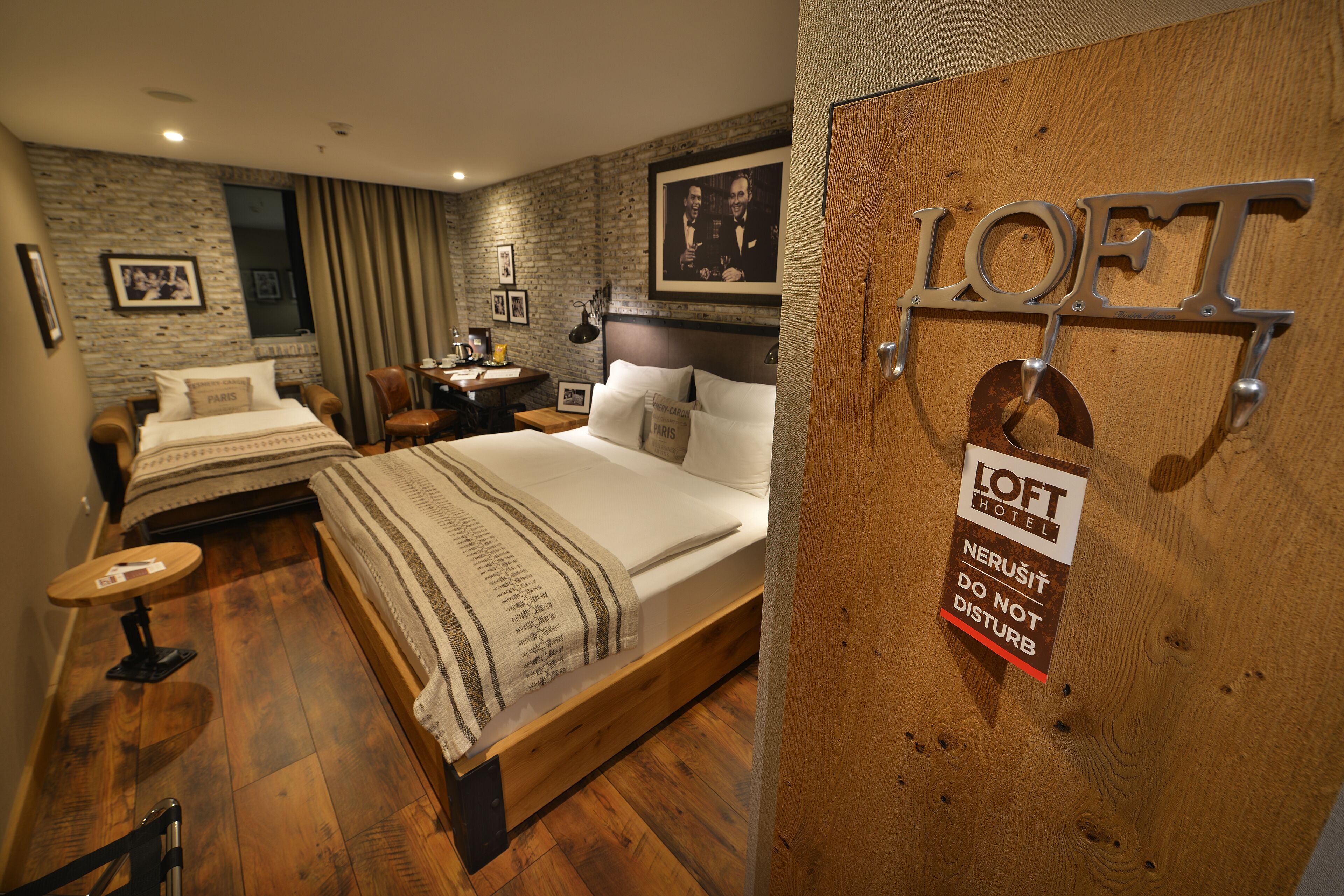 A hotel room with a bed, a desk, a chair, and a small table. There is a sign on the door that reads "LOFT HOTEL" and "NEURUSIT DO NOT DISTURB".