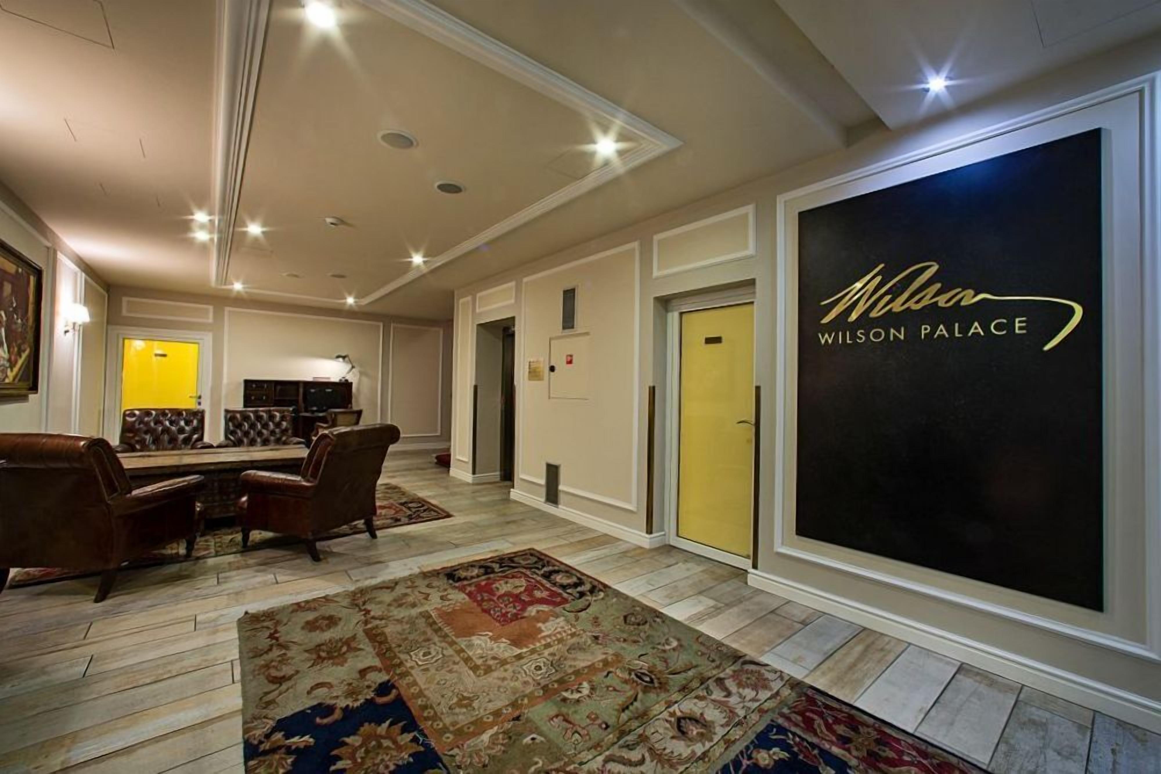 En rymlig hotellobby med en stor, svart skylt med texten ”Wilson Palace”, en matplats med bruna läderstolar och en mönstrad matta på golvet.